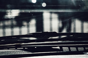 The image captures a close-up view of a car windshield and dashboard. The focus is on the windshield wipers with a blurred background showing some light reflections. The image has a dark and shadowy ambiance, with some light filtering through.