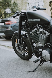 A close-up view of a Harley-Davidson motorcycle parked on a street. The motorcycle's shiny engine and exhaust pipes are prominently displayed, highlighting its powerful build. A car is parked in the background, partially visible.