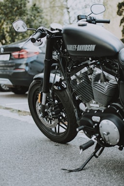 A close-up view of a Harley-Davidson motorcycle parked on a street. The motorcycle's shiny engine and exhaust pipes are prominently displayed, highlighting its powerful build. A car is parked in the background, partially visible.