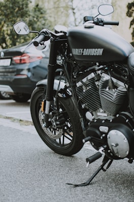 A close-up view of a Harley-Davidson motorcycle parked on a street. The motorcycle's shiny engine and exhaust pipes are prominently displayed, highlighting its powerful build. A car is parked in the background, partially visible.