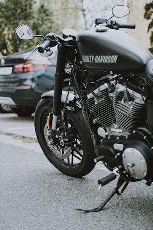 A close-up view of a Harley-Davidson motorcycle parked on a street. The motorcycle's shiny engine and exhaust pipes are prominently displayed, highlighting its powerful build. A car is parked in the background, partially visible.