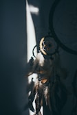 A dreamcatcher hangs in shadowed light, with its circular web accented by feathers and beads. The play of light creates a moody and mystical atmosphere.