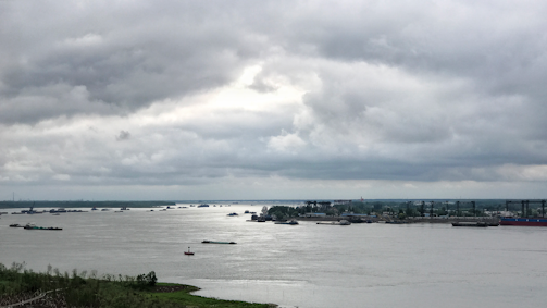A wide river is filled with multiple cargo ships and barges under a cloudy sky. The horizon is dominated by industrial structures and cranes, indicating a port area. The water appears calm, and a small portion of greenery can be seen in the foreground.