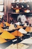 A variety of colorful spices are displayed in conical piles on a market stall, each labeled with small black signs. In the background, large jars filled with different substances are neatly arranged on shelves.