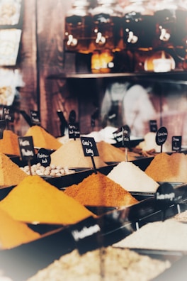 A variety of colorful spices are displayed in conical piles on a market stall, each labeled with small black signs. In the background, large jars filled with different substances are neatly arranged on shelves.