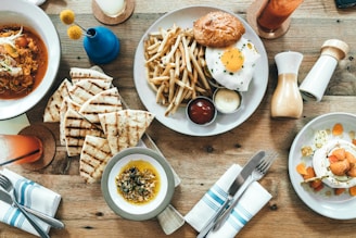 A wooden table is set with an assortment of dishes and items, including a burger with an egg on top and fries, a bowl of soup or stew with grilled bread, a plate with a creamy dessert topped with fruit, and small bowls with sauces. There are also condiments like salt and pepper shakers, a blue vase with flowers, and drinks in tall glasses.