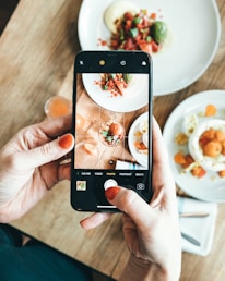 A person is holding a smartphone and capturing a photo of a plated dish on a wooden table. The dish appears to be a gourmet meal with various colorful ingredients, including green, red, and orange elements. There is another plate and a small glass in the background.