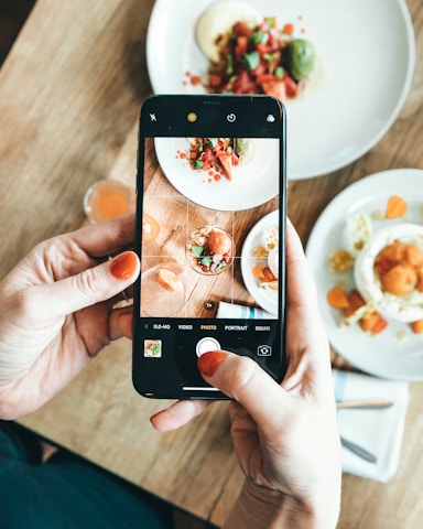 A person is holding a smartphone and capturing a photo of a plated dish on a wooden table. The dish appears to be a gourmet meal with various colorful ingredients, including green, red, and orange elements. There is another plate and a small glass in the background.