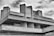 A Brutalist architectural structure with distinct geometric shapes and exposed concrete surfaces. The building features angular lines and large, rectangular columns, set against a partly cloudy sky.
