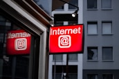 A brightly lit red sign displays the word 'Internet' with the @ symbol, mounted outside a building with a modern facade and several windows.
