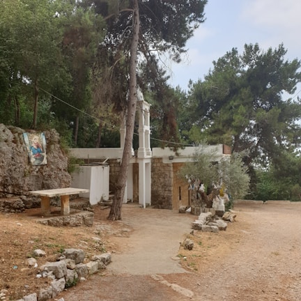 A tranquil outdoor scene featuring a small chapel built with stone and plaster, surrounded by lush green trees. A pathway made of dirt and stones leads to the entrance. To the left, there is a religious painting mounted on a rock and a stone table nearby, suggesting a place for reflection or gathering. Piles of rocks and a statue of religious figures can be seen under the trees on the right, adding to the serene atmosphere.