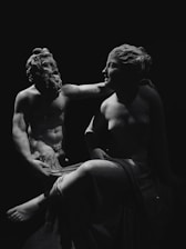 Two classical sculptures are captured in a black and white photo, featuring a male and female figure. The male figure has a beard and a muscular build, with a gentle expression, and appears to be playfully interacting with the female figure. The female figure is seated, with a serene expression, and is partially draped in fabric. Both sculptures are highlighted dramatically against a dark background, emphasizing their contours and forms.