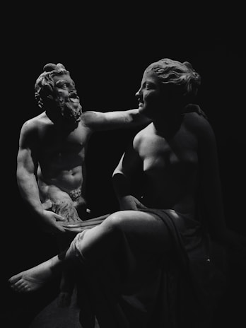 Two classical sculptures are captured in a black and white photo, featuring a male and female figure. The male figure has a beard and a muscular build, with a gentle expression, and appears to be playfully interacting with the female figure. The female figure is seated, with a serene expression, and is partially draped in fabric. Both sculptures are highlighted dramatically against a dark background, emphasizing their contours and forms.