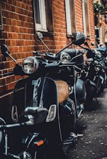 A row of black scooters parked along a brick wall, with reflections visible on the scooter surfaces. The setting appears to be urban, with narrow space between the vehicles and the building. The scooters have a retro design with round mirrors and headlights.