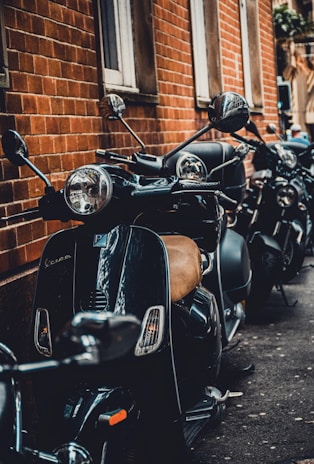 A row of black scooters parked along a brick wall, with reflections visible on the scooter surfaces. The setting appears to be urban, with narrow space between the vehicles and the building. The scooters have a retro design with round mirrors and headlights.