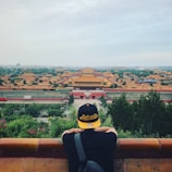 A person wearing a black and yellow cap with the word 'Authentic' is standing with their back to the camera, overlooking a vast historical site with traditional Chinese architecture, featuring numerous yellow-roofed buildings amidst greenery and a cloudy sky.