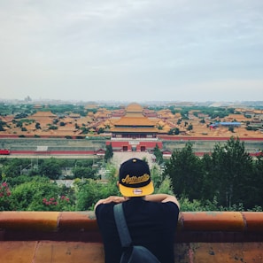 A person wearing a black and yellow cap with the word 'Authentic' is standing with their back to the camera, overlooking a vast historical site with traditional Chinese architecture, featuring numerous yellow-roofed buildings amidst greenery and a cloudy sky.