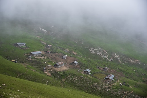 A series of small, rustic cabins are scattered across a lush green hillside enveloped in mist. The cabins have simple, metal rooftops and are connected by dirt paths winding through the vibrant landscape. The mist creates a serene and mysterious atmosphere, partially obscuring the distant terrain.