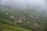 A series of small, rustic cabins are scattered across a lush green hillside enveloped in mist. The cabins have simple, metal rooftops and are connected by dirt paths winding through the vibrant landscape. The mist creates a serene and mysterious atmosphere, partially obscuring the distant terrain.