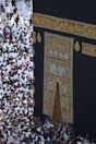 A large gathering of people surrounds the Kaaba, a sacred Islamic structure covered with an ornate black and gold cloth featuring Arabic calligraphy. People are dressed mostly in white robes, which is traditional attire for pilgrims.