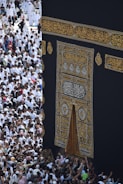 A large gathering of people surrounds the Kaaba, a sacred Islamic structure covered with an ornate black and gold cloth featuring Arabic calligraphy. People are dressed mostly in white robes, which is traditional attire for pilgrims.