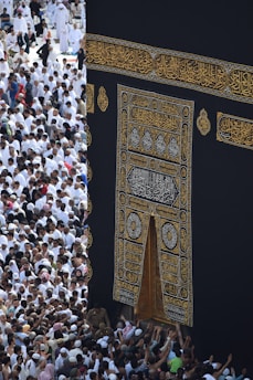 A large gathering of people surrounds the Kaaba, a sacred Islamic structure covered with an ornate black and gold cloth featuring Arabic calligraphy. People are dressed mostly in white robes, which is traditional attire for pilgrims.