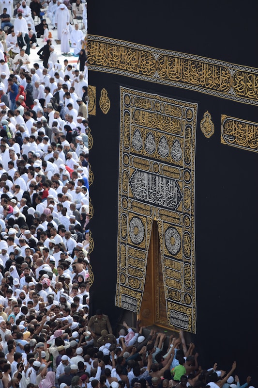 A large gathering of people surrounds the Kaaba, a sacred Islamic structure covered with an ornate black and gold cloth featuring Arabic calligraphy. People are dressed mostly in white robes, which is traditional attire for pilgrims.