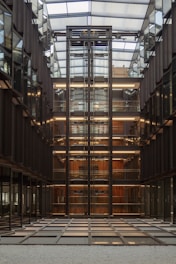 A modern interior features a central glass elevator shaft with exposed metal beams, surrounded by multiple floors of office spaces. The design includes symmetrical lines and reflective surfaces, highlighting a sense of order and precision. The ceiling is glass, allowing natural light to illuminate the space.