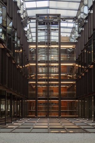 A modern interior features a central glass elevator shaft with exposed metal beams, surrounded by multiple floors of office spaces. The design includes symmetrical lines and reflective surfaces, highlighting a sense of order and precision. The ceiling is glass, allowing natural light to illuminate the space.