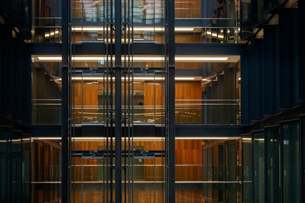 Several floors of an office building are visible through large glass walls. The interior features a modern, industrial design with exposed structural elements and wooden paneling. Horizontal lighting fixtures illuminate the space along the corridors.