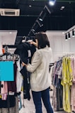 A woman is shopping for clothes in a store. She is holding a black garment on a hanger while looking at it. The store has several racks filled with a variety of clothes including shirts, blouses, and jackets. The setting is well-lit with ceiling lights focused on the clothes. An air conditioning vent is visible on the ceiling.