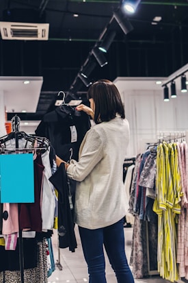 A woman is shopping for clothes in a store. She is holding a black garment on a hanger while looking at it. The store has several racks filled with a variety of clothes including shirts, blouses, and jackets. The setting is well-lit with ceiling lights focused on the clothes. An air conditioning vent is visible on the ceiling.