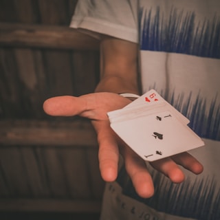 A person extends their hand, holding a fanned-out deck of playing cards with the ace and seven of spades visible. The shirt worn by the person features a blue graphic design, and the background is a wooden surface.