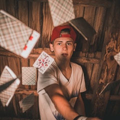 A person wearing a red cap is surrounded by playing cards that are mid-air, suggesting motion. The scene is set against a wooden backdrop, and the person is reaching out with one arm, possibly to throw or catch the cards.