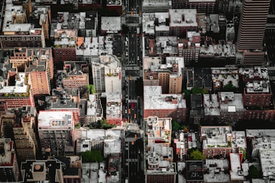 An aerial view of an urban landscape, displaying numerous high-rise buildings with diverse architectural styles, arranged in a grid pattern. The rooftops of these buildings are varying shades of white and gray, some of which have greenery or HVAC equipment. A central road divides the buildings, with visible traffic and crosswalks. Streets and intersections are lined with trees, providing patches of green among the concrete.
