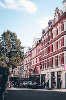Red brick buildings with white accents line a city street under a blue sky. Several people are walking along the sidewalk, and bicycles are parked nearby. There are trees and cars along the road, creating a lively urban environment.