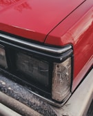 A close-up of the front part of a red vehicle, focusing on the headlight and part of the bumper. The surface of the car appears to be slightly wet, suggesting recent rain or washing. The headlight design is rectangular and surrounds the corner area along with other lights.