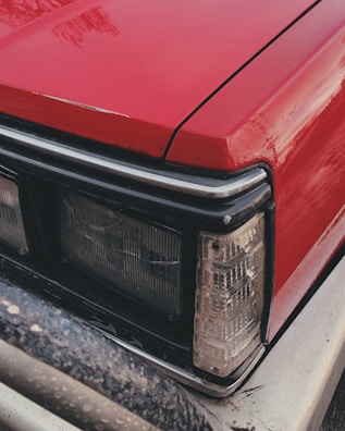 A close-up of the front part of a red vehicle, focusing on the headlight and part of the bumper. The surface of the car appears to be slightly wet, suggesting recent rain or washing. The headlight design is rectangular and surrounds the corner area along with other lights.