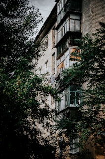 A multi-story residential building with brick exterior partially obscured by lush green trees. The building features a number of enclosed balconies with glass panes and some visible laundry. The overall structure seems aged, with weathered textures.