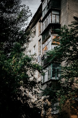 A multi-story residential building with brick exterior partially obscured by lush green trees. The building features a number of enclosed balconies with glass panes and some visible laundry. The overall structure seems aged, with weathered textures.