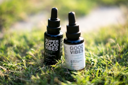 Two dropper bottles labeled 'Super Good Vibes' and 'Good Vibes' sit on a patch of green grass. The bottles are dark with black caps and white labeling containing small text. Sunlight illuminates the grass, creating a warm and relaxed atmosphere.