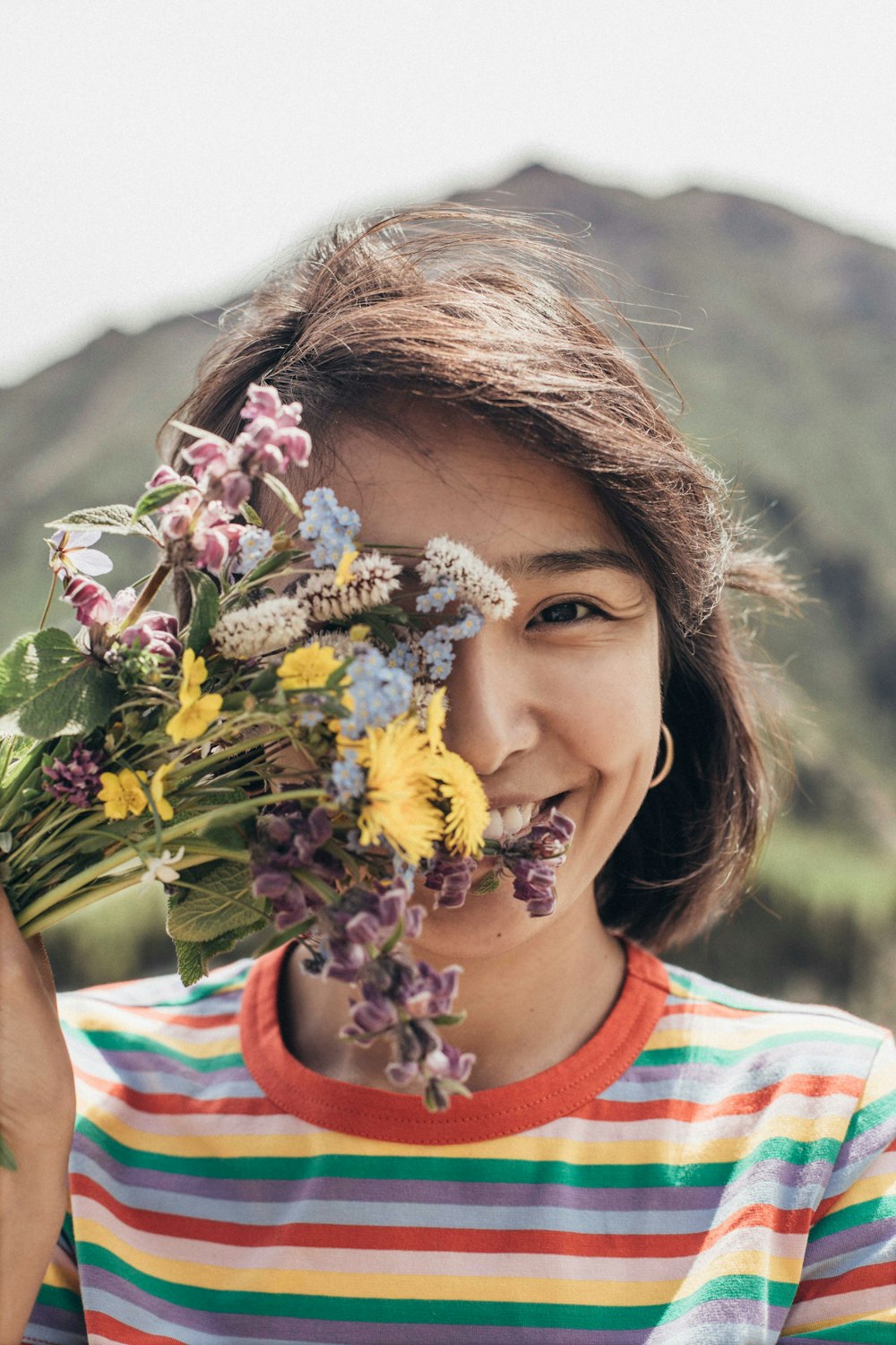 girl standing and covering her right eye with petaled flowers and smiling