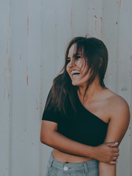 A person with long hair is laughing joyfully, wearing a black off-shoulder top and light blue jeans. The background consists of an off-white wall with subtle lines and minimal texture.