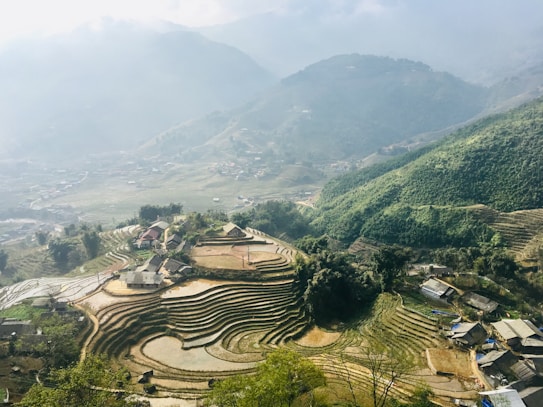 A picturesque mountainous landscape featuring terraced rice fields. The terraced fields gradually ascend the mountainside, surrounded by dense green vegetation. A few scattered traditional houses are visible near the fields, adding a sense of rural charm. Mist shrouds the distant hills, creating a serene and tranquil atmosphere.