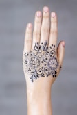 A hand with intricate henna art on the top, featuring floral motifs and swirling patterns. The design is symmetrical and detailed, focusing on two main flower shapes surrounded by leaves and dots.