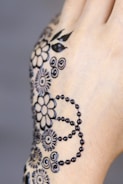 Intricate black henna patterns are delicately designed on the back of a hand. The design features floral motifs, circular patterns, and dotted lines, creating an elegant and artistic effect.