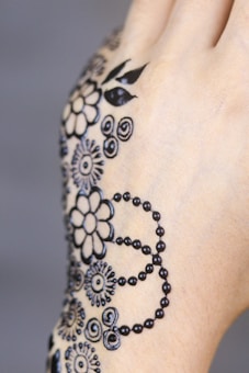 Intricate black henna patterns are delicately designed on the back of a hand. The design features floral motifs, circular patterns, and dotted lines, creating an elegant and artistic effect.