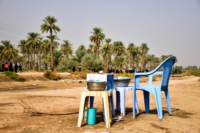 A rural scene features a couple of blue plastic chairs and a small white plastic table on a dirt surface. On the table, there are a few metal bowls and a green jug. In the background, a grove of tall palm trees can be seen, and a few people are walking by.