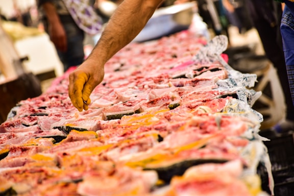 A hand reaches over a market stall displaying a wide array of fresh fish. The fish are sliced open, revealing their pink and white flesh against a backdrop of market activity.