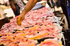 A hand reaches over a market stall displaying a wide array of fresh fish. The fish are sliced open, revealing their pink and white flesh against a backdrop of market activity.